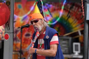 Merry Prankster George Walker plays a kazoo taped to an axe in front of a multicolored psychedelic painted backdrop.