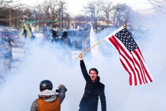A counterprotestor holds up an American flag as tear gas dispersed by federal agents behind him in Saint Paul, Minnesota, on January 8, 2026. (Credit: CHARLY TRIBALLEAU / AFP via Getty Images)